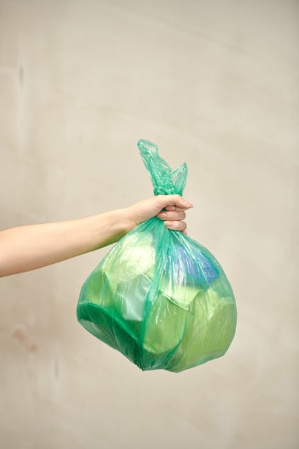 A person's arm extends into the frame from the left, holding a green and blue plastic rubbish bag by its tied-off top with their hand. The bag appears full and somewhat rounded, with a textured surfac